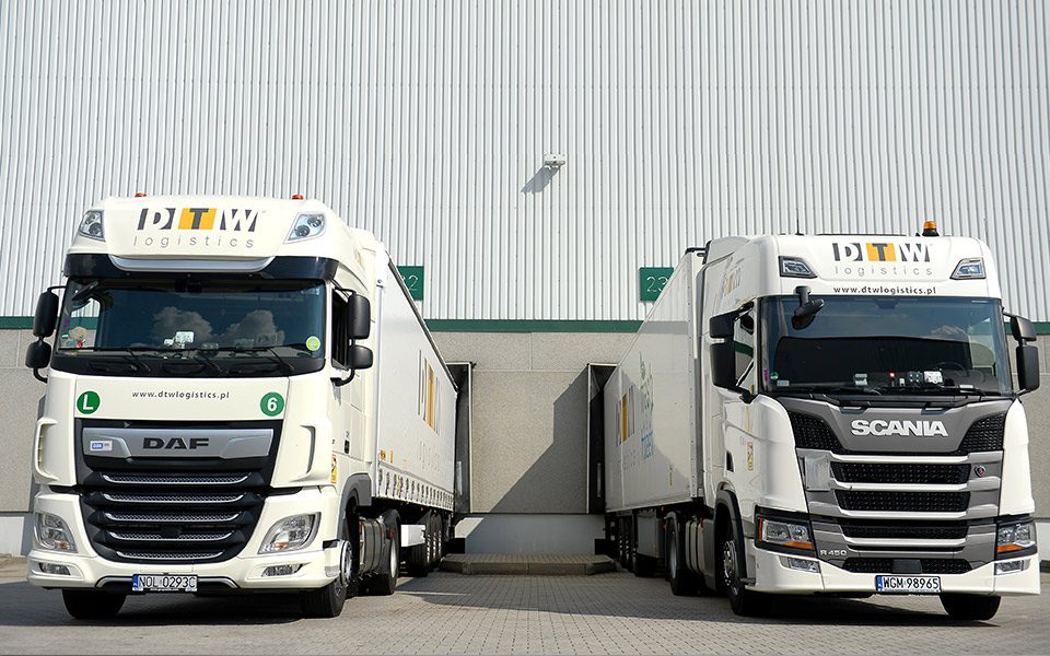 Dwie ciężarówki DTW Logistics Group stojące przed magazynem podczas załadunki i rozładunku.